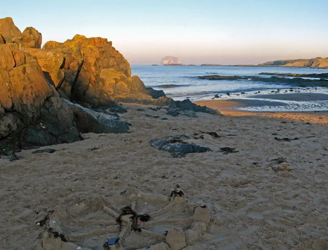 Rocky sandy beach at sunset with golden boulders, calm sea, and distant headlands under clear sky.