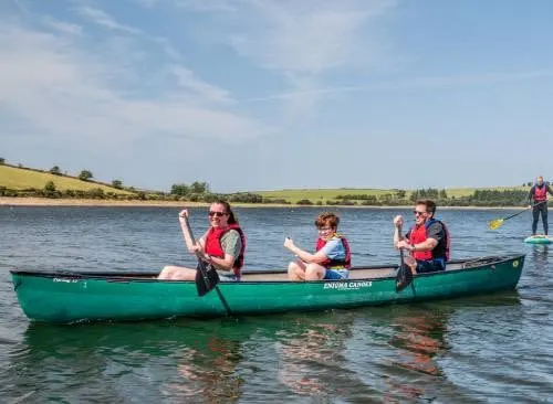 A group of children in red life jackets paddle a green canoe on a calm lake with green fields and trees visible in the background.