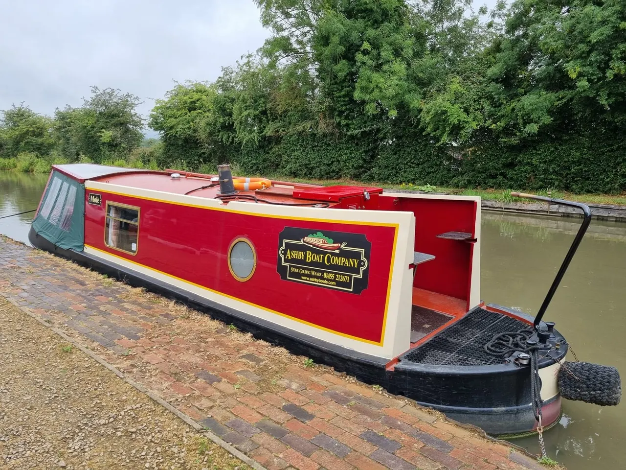Red and cream narrowboat moored on a canal with tree-lined banks in the background.