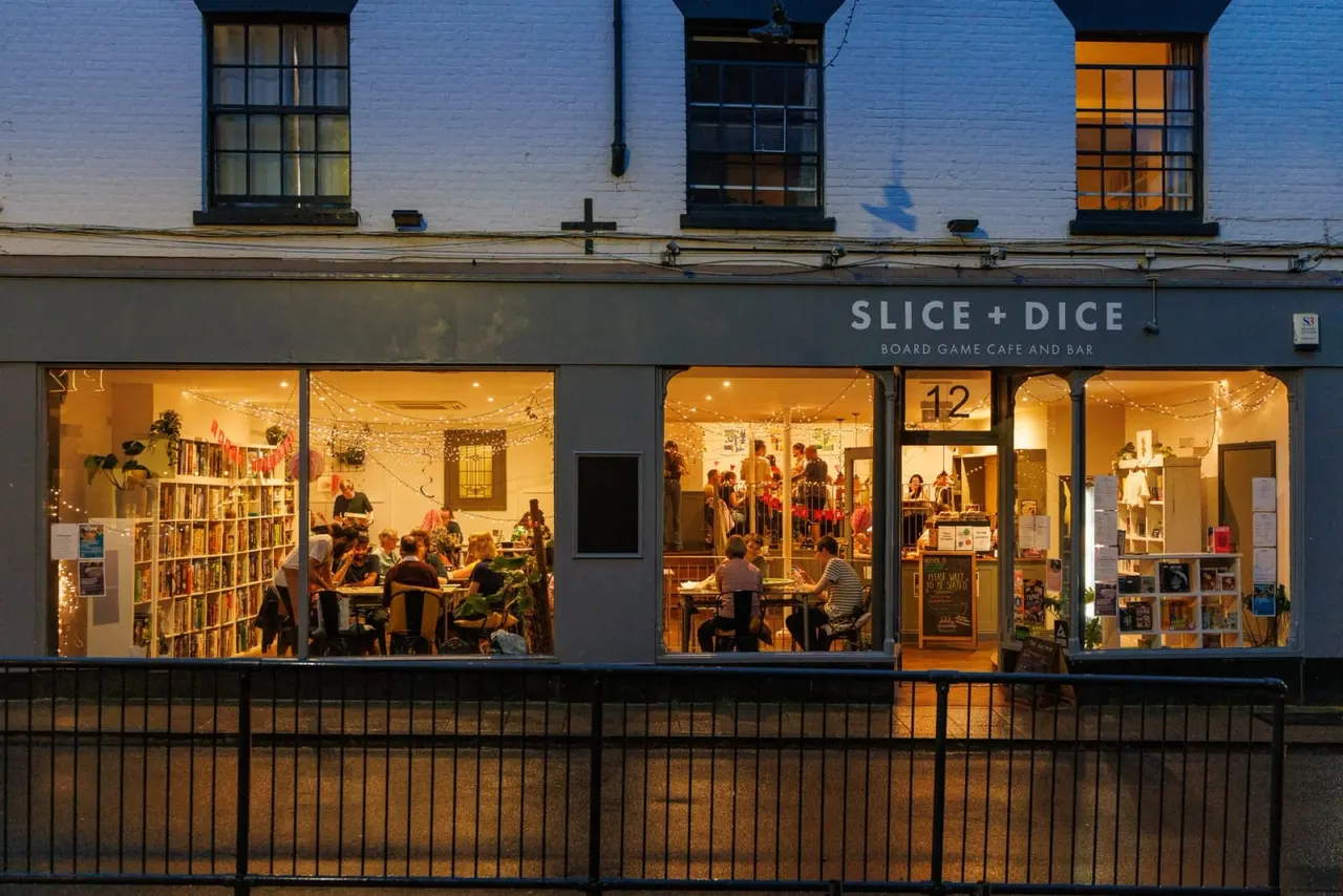 Illuminated storefront of a board game café and bar with multiple customers seated at tables inside, visible through large windows.