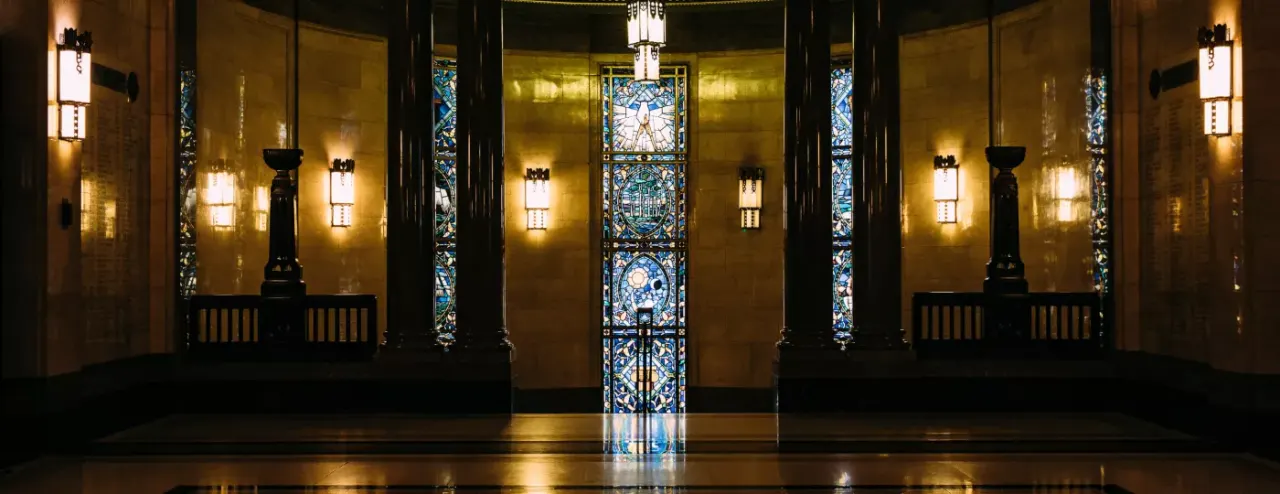 Symmetrical Art Deco interior with golden walls, ornate stained-glass doors, black pillars, and wall-mounted lighting fixtures.