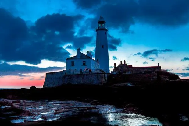 Lighthouse and keeper's cottage on rocky shore at twilight with dramatic blue-toned sky.