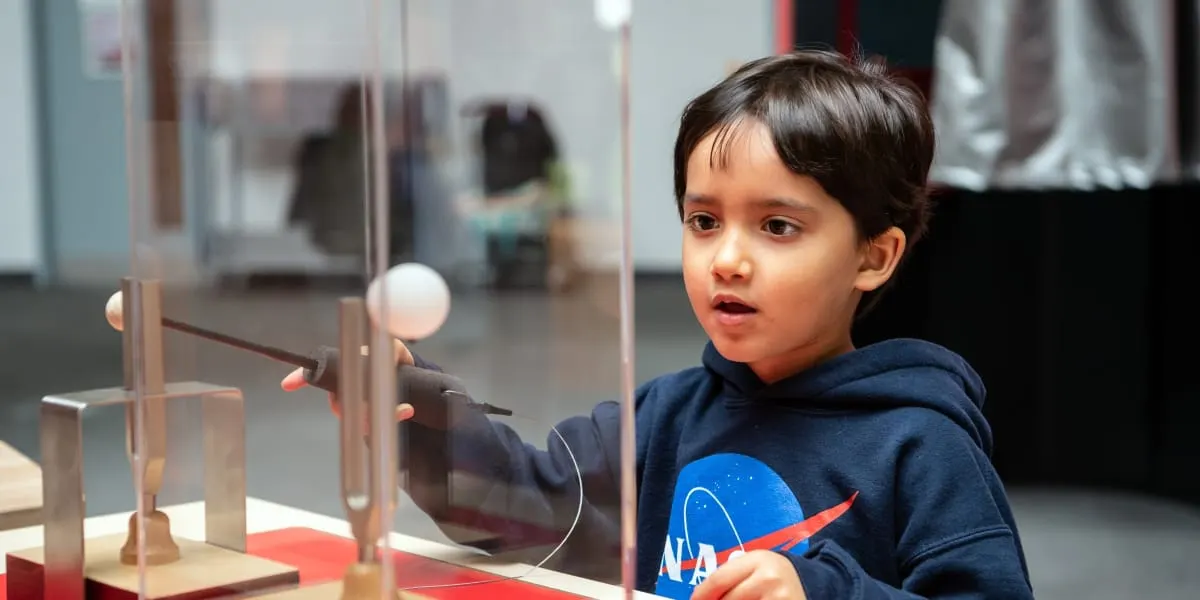 Child in blue NASA hoodie interacts with a hands-on science exhibit featuring spheres and metal rods.