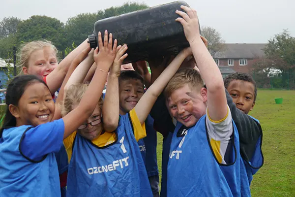 A group of children in blue sports shirts lifting a large black tyre together outdoors.