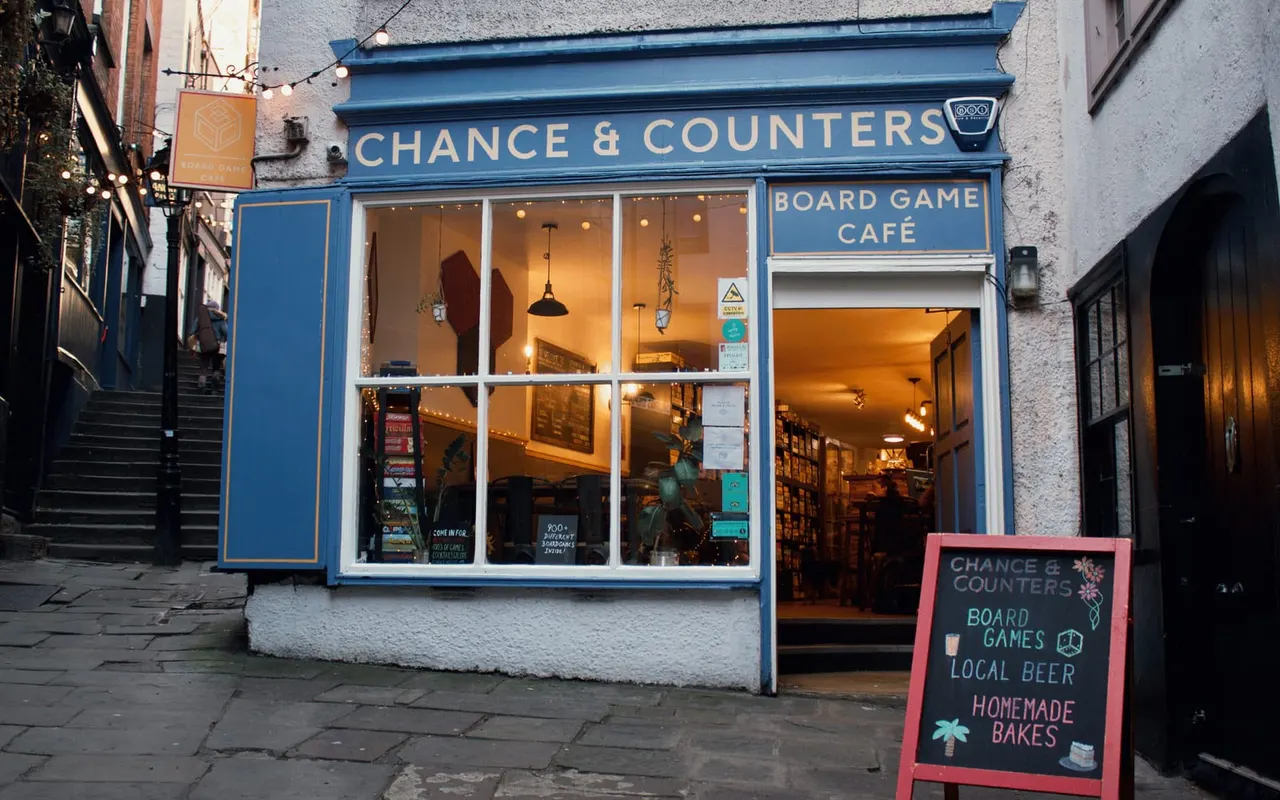 Front exterior of Chance & Counters board game café on Christmas Steps, with blue signage, illuminated interior, and a pink sandwich board advertising games, beer and homemade bakes.