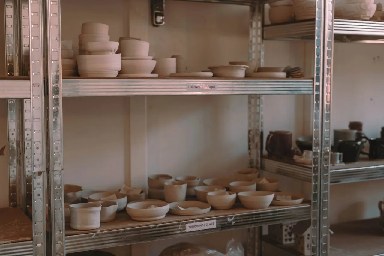 Shelves displaying unfired ceramic bowls and vessels in various sizes, arranged on metal industrial shelving in a pottery studio.
