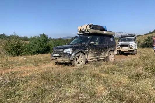 Two rugged 4x4 vehicles with roof racks parked in grassy countryside terrain under clear blue sky.