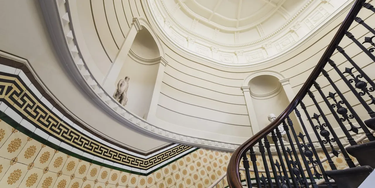 Looking up at a grand curved ceiling with decorative plasterwork, classical sculptures and ornate wrought-iron staircase railings.