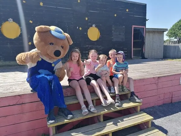 Five children sit on wooden steps next to a large teddy bear mascot wearing blue, with a themed building behind them.