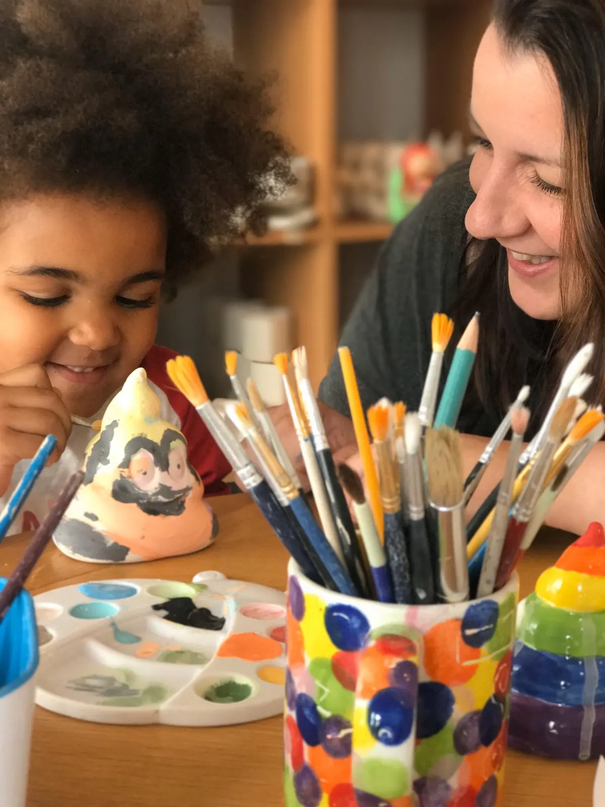 Adult and child doing arts and crafts together at a table with paintbrushes, paints and a decorated pottery piece.