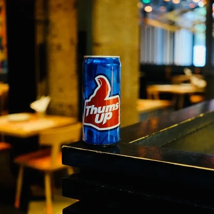 A can of Thumbs Up soft drink sits on a dark counter in a dimly lit venue with warm lighting and blurred interior details in the background.