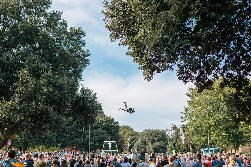 Person performing an acrobatic flip in mid-air above a trapeze rig, watched by a large crowd in a tree-lined park.