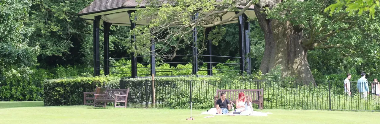 Two people picnicking on grass in a park with mature trees, pergola and benches visible behind a fence.