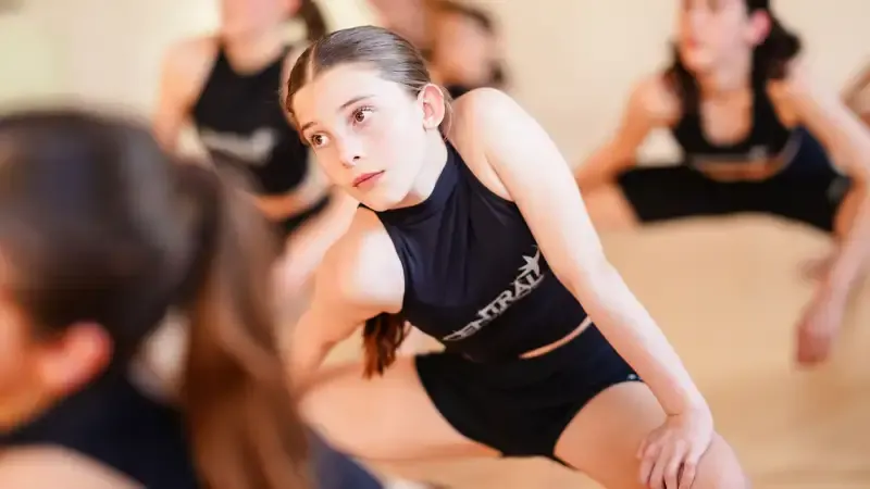 Young dancer in black sportswear performing in a studio with other participants in the background.