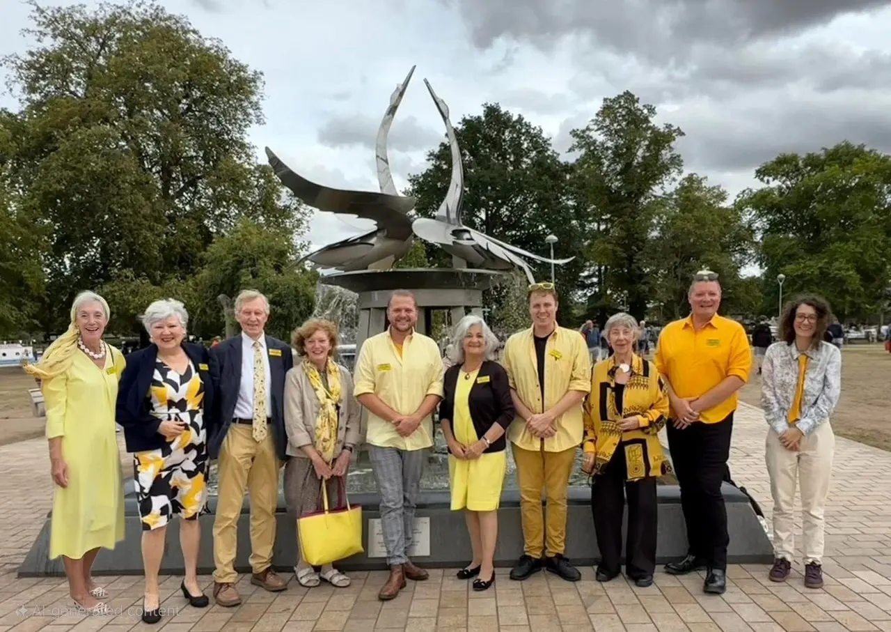 Group of ten people in yellow and navy clothing standing in front of a large metal sculpture with curved horns in a park setting.