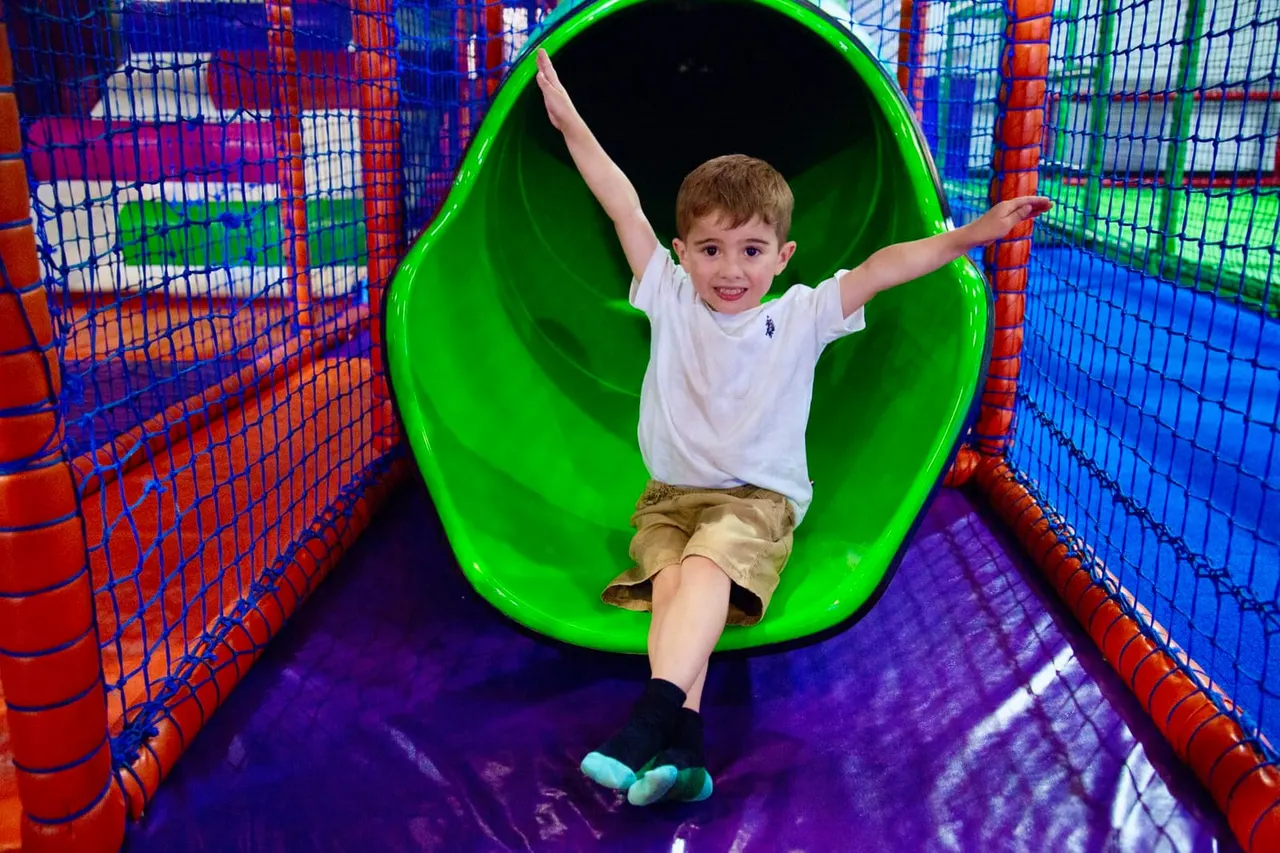 Young child sliding down a bright green chute with arms outstretched inside a colourful soft play structure.