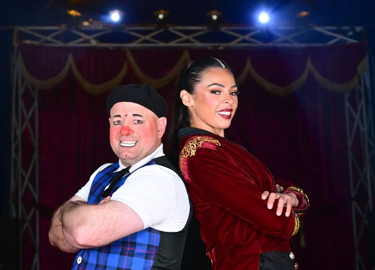 A clown in red nose and tartan trousers poses with a woman in a red velvet jacket on a circus stage with draped curtains and stage lights.