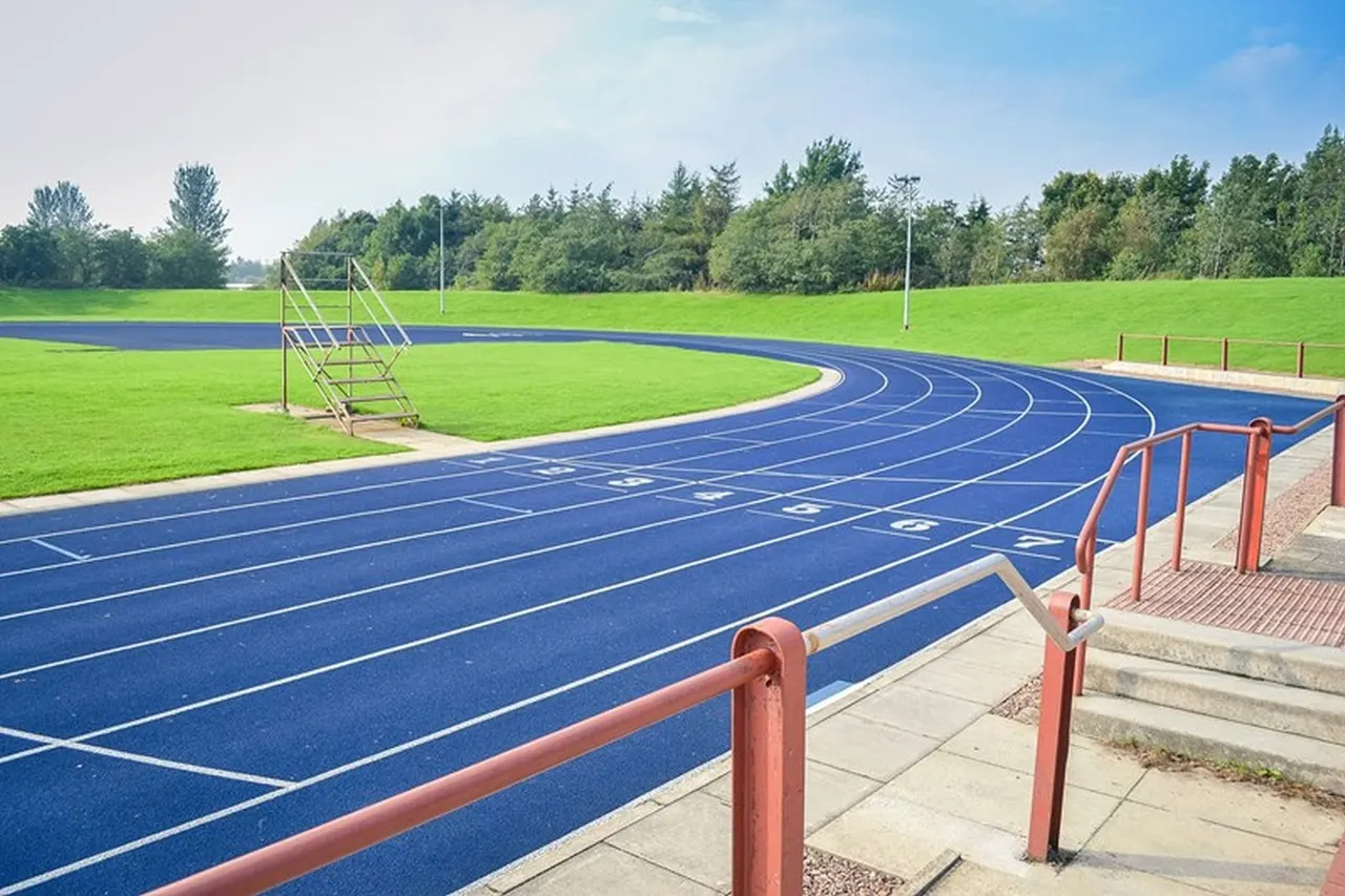Blue running track with numbered lanes at an outdoor sports facility, with green field and trees beyond.