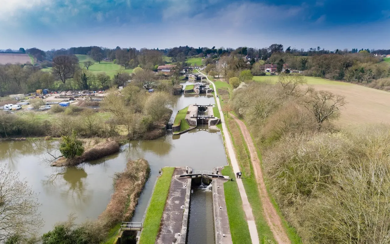 Aerial view of a canal with multiple lock gates stretching into the distance, surrounded by green fields, scattered trees, and rural buildings.