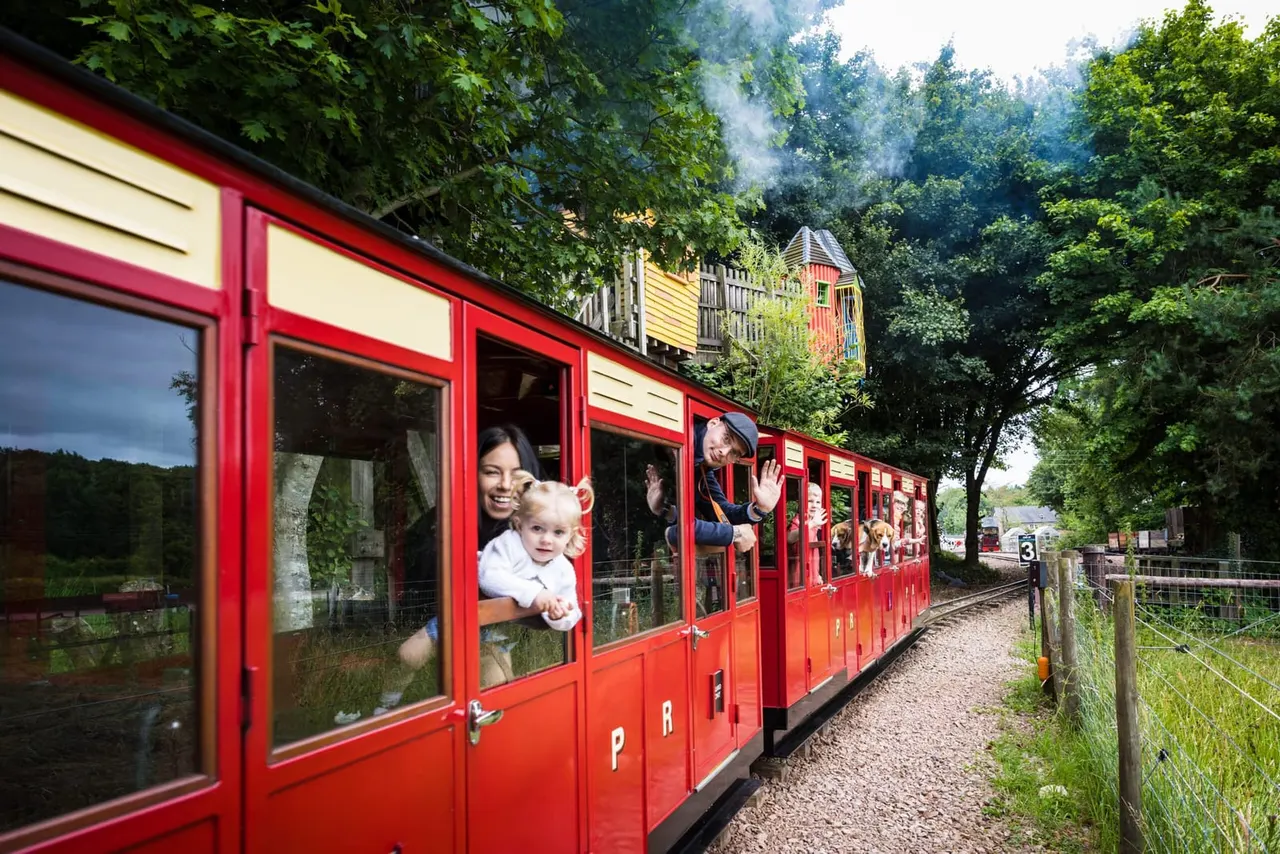 Families and children riding in red open-sided carriages on a heritage railway surrounded by woodlands and colourful station buildings.