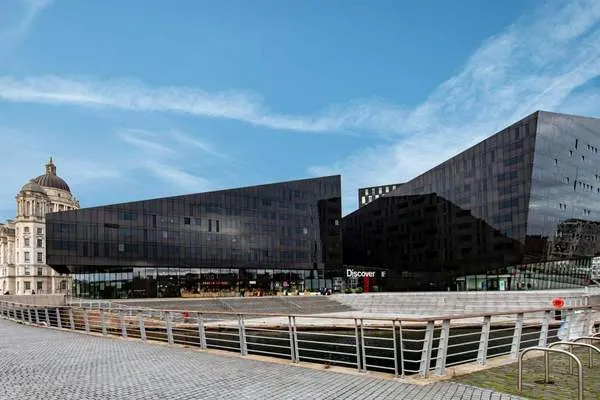 Modern black building with angular architecture beside historic dome, waterfront plaza with metal railings in foreground.