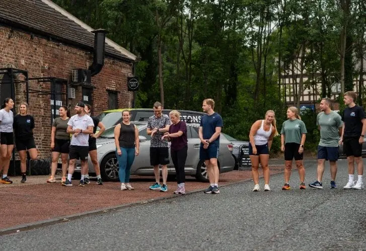 A group of people in athletic wear gathered outside a brick building before or after a fitness session.