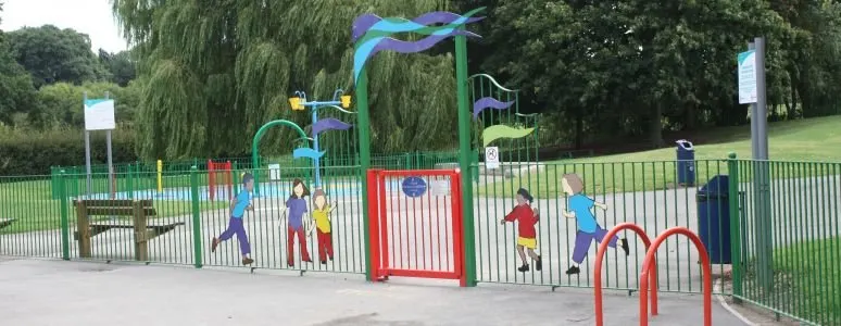 Children playing at a colourful playground with climbing frames, slides and archways surrounded by fencing and trees.