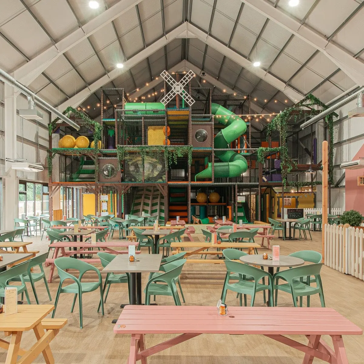 Indoor play barn with dining area featuring green chairs and tables, large multi-level play structure with slides and climbing equipment in background.