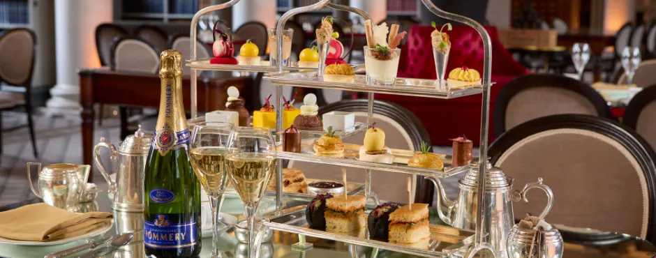Tiered glass stand displaying an afternoon tea service with pastries, sandwiches and cakes, champagne bottle and glasses on dining table.