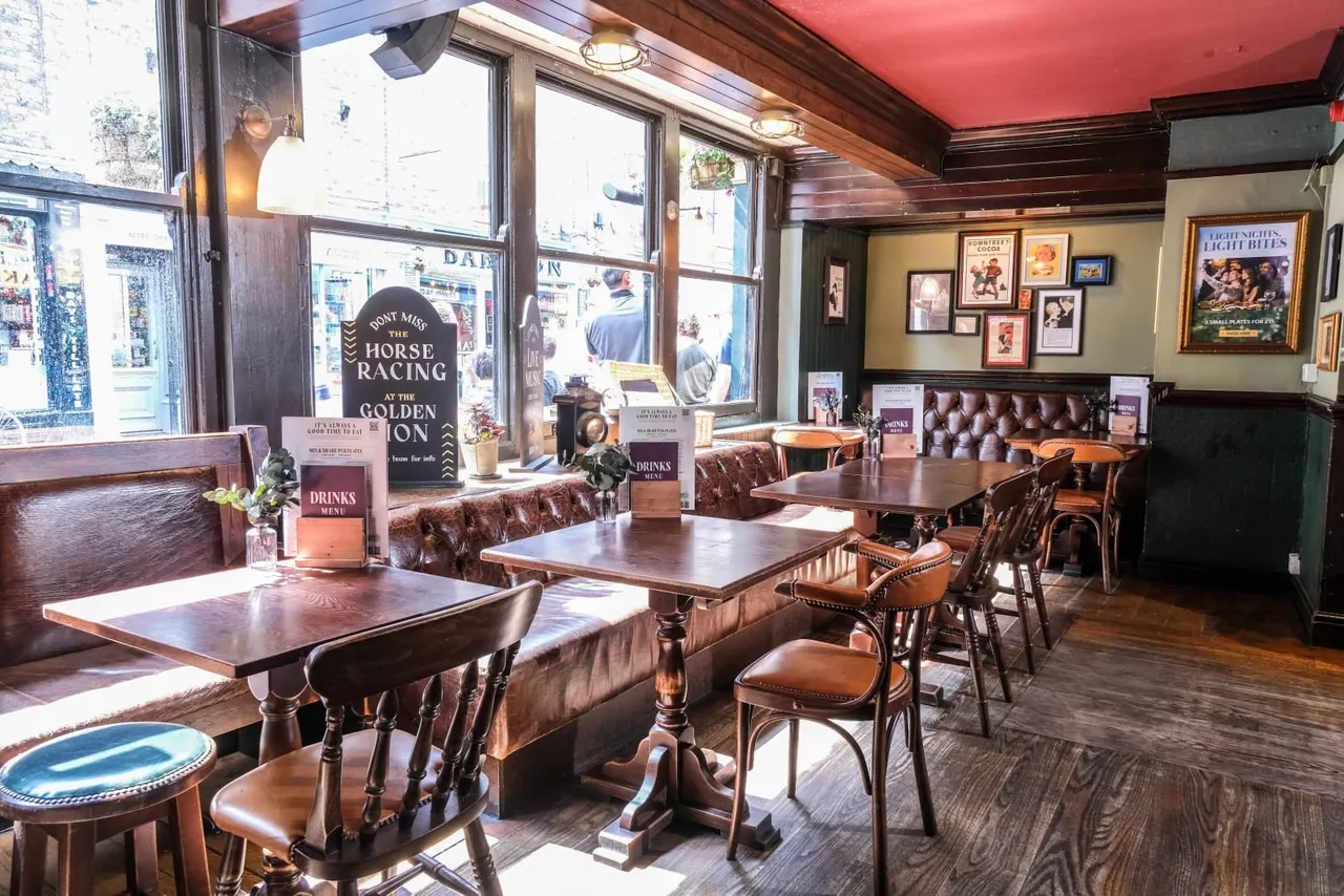 Interior of a traditional pub with wooden tables, leather bench seating, framed horse racing posters on walls, and large street-facing windows.