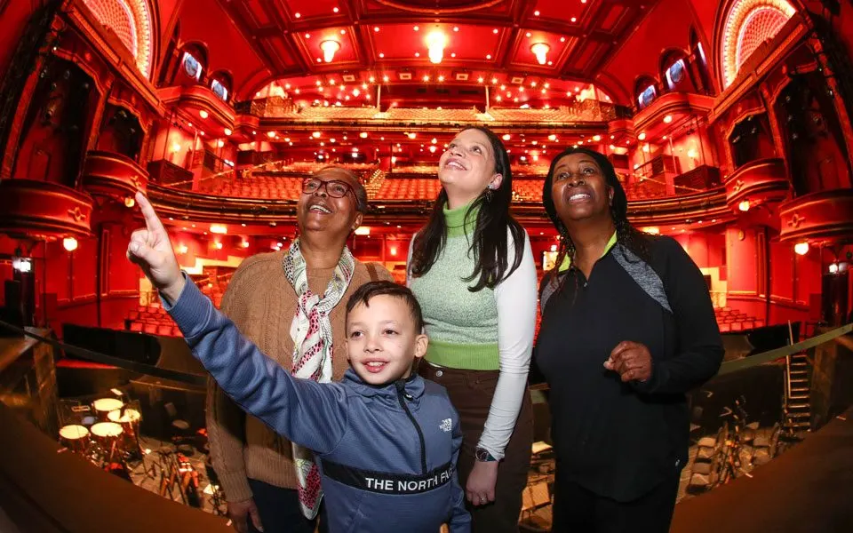 Four people, including a young child, stand inside an ornate theatre auditorium lit in red, gazing upward at the illuminated balconies and ceiling.