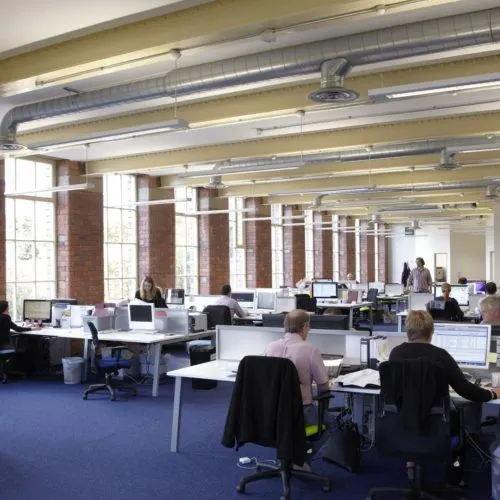 Open-plan office space with multiple workers at desks with computers, industrial ceiling, and brick columns alongside large windows.