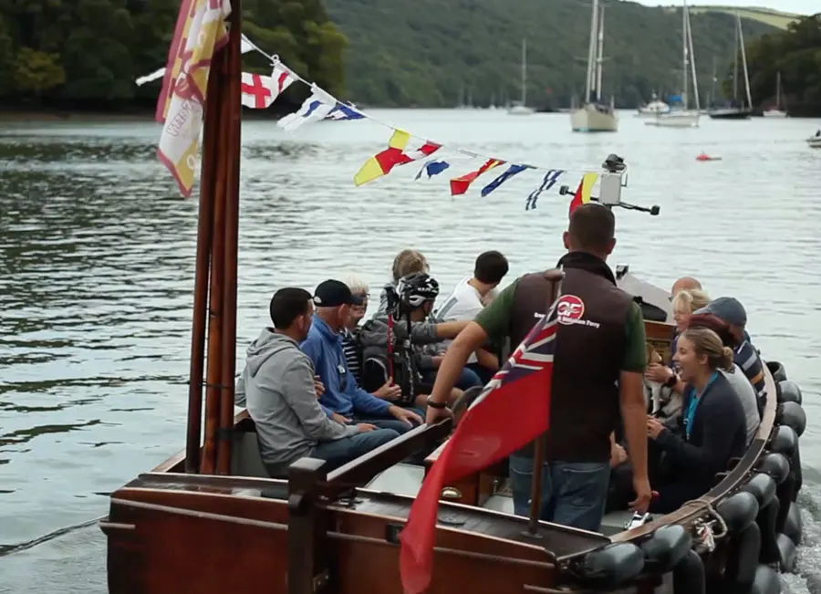 Passengers board a traditional wooden ferry boat at a riverside dock, with sailing boats visible on the water behind.
