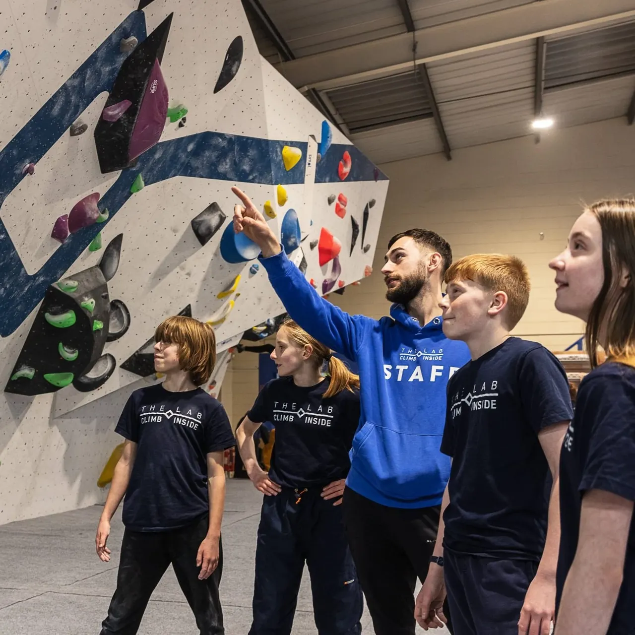 An instructor in blue points out climbing holds to a group of children wearing matching black t-shirts at an indoor climbing wall.