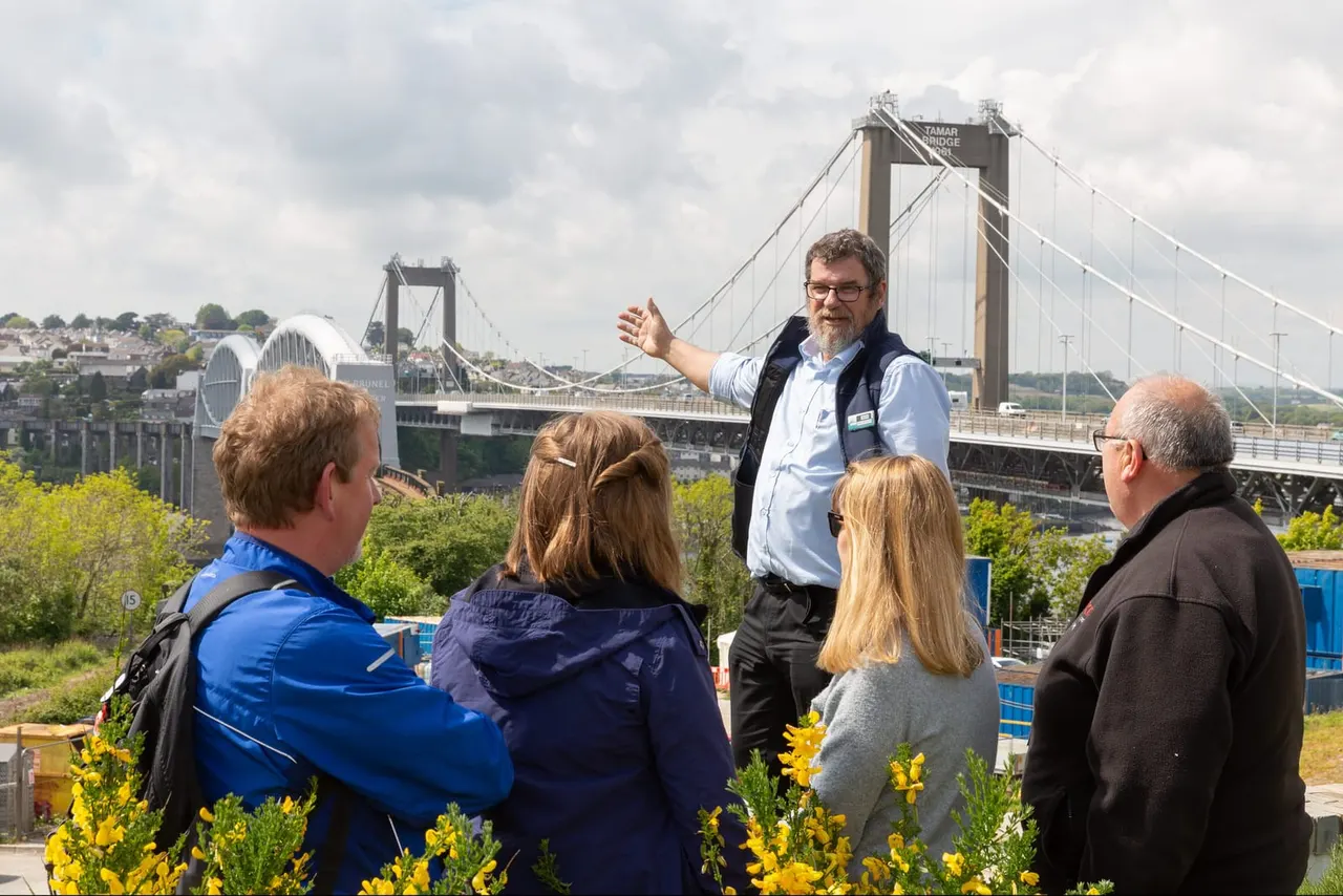 A tour guide gestures while speaking to a group of visitors overlooking the Tamar Bridge from a hillside viewpoint.