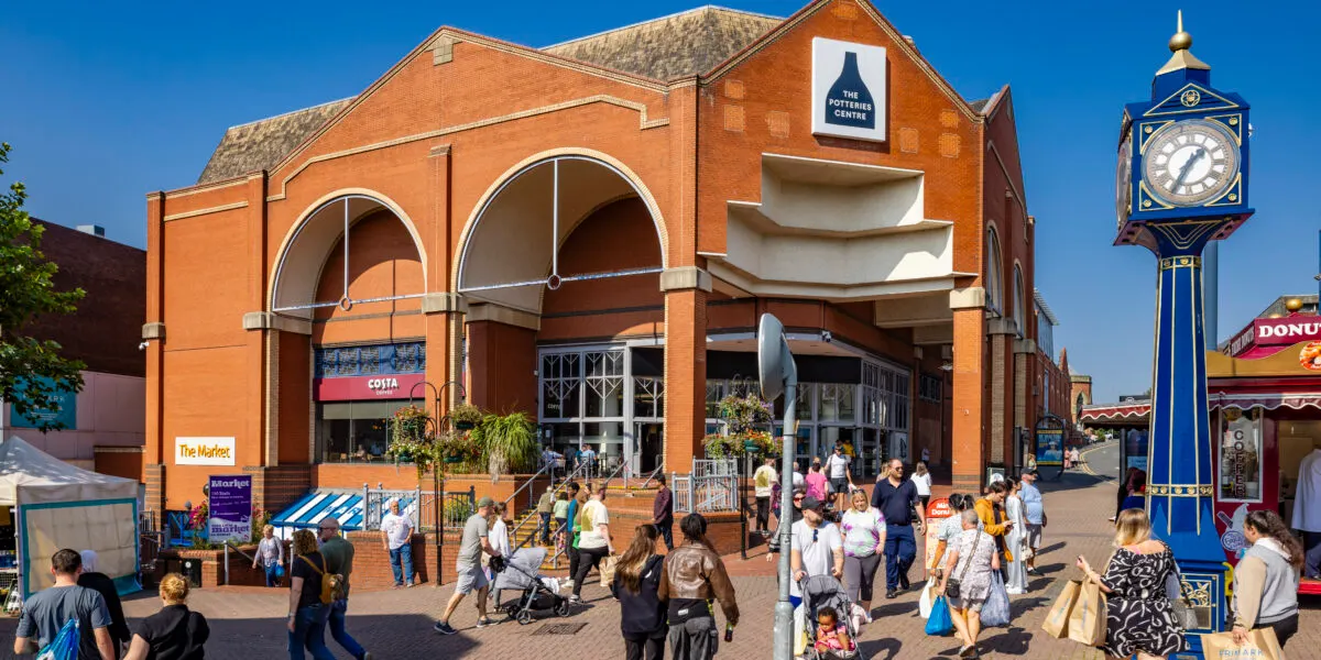 Busy outdoor shopping area with visitors walking past a brick market building, clock tower and retail outlets on a clear day.