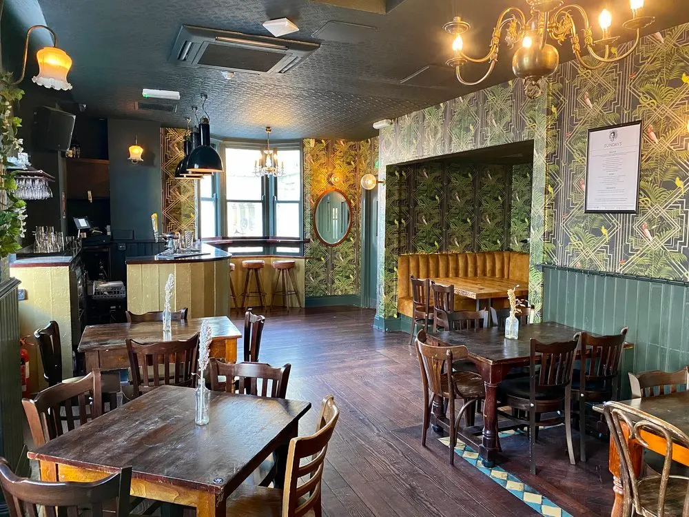 Interior of a gastropub with wooden dining tables and chairs, a bar area with stools, and decorative patterned wallpaper and brass lighting fixtures.