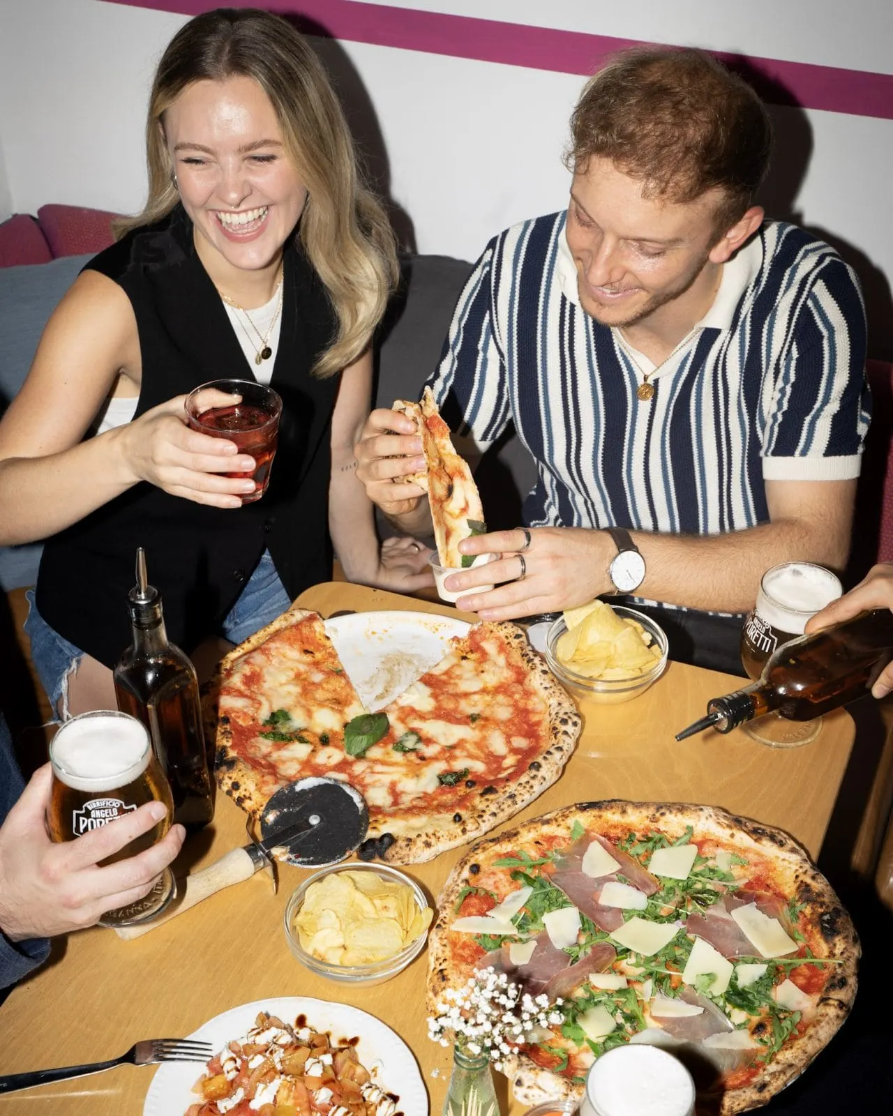 Two people eating pizza and sharing drinks at a table with multiple pizzas and side dishes.