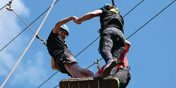 Two people on a high ropes course, one climbing a wooden pole whilst tethered with safety equipment against a blue sky.