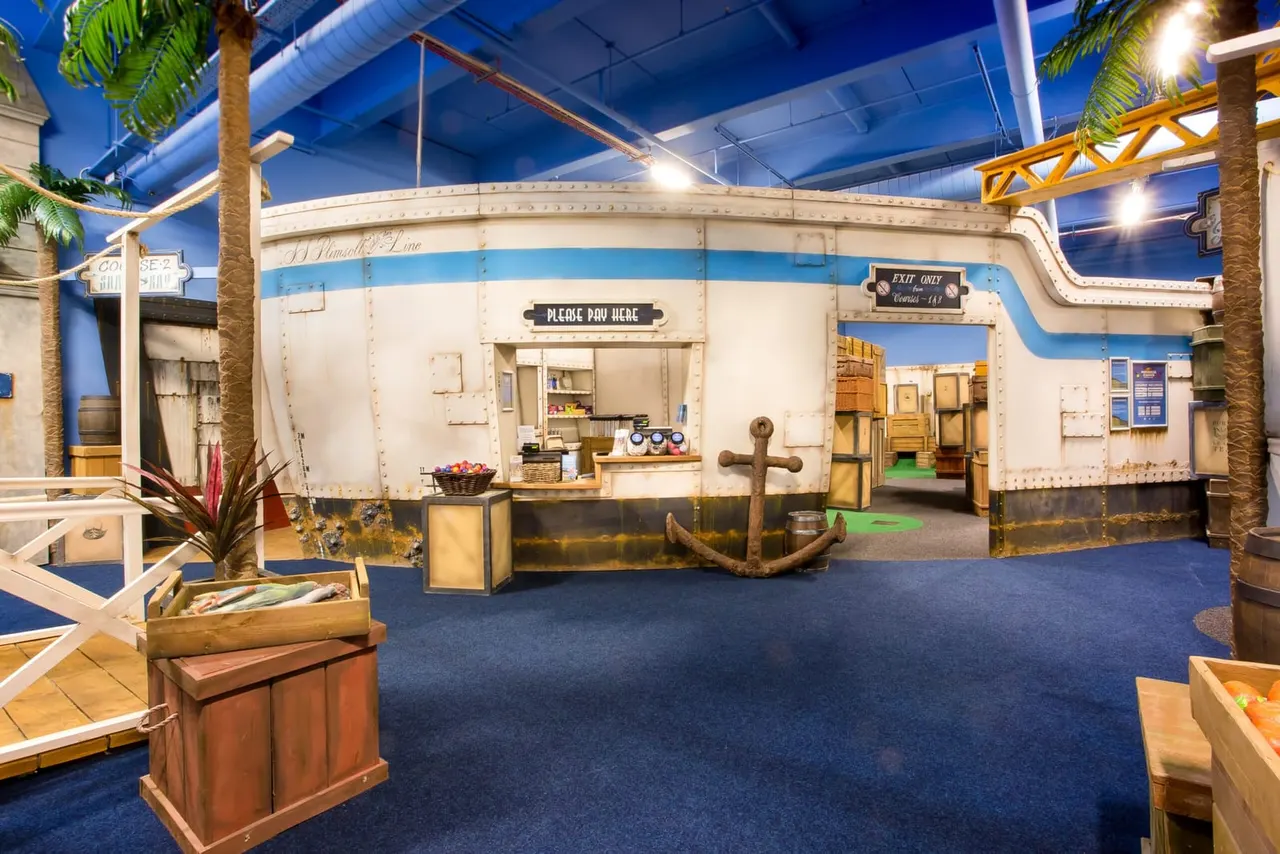 Indoor adventure golf course with vintage caravan-themed décor, nautical props and palm trees under blue ceiling.