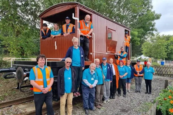 Group of volunteers and staff in blue and orange uniforms posing by a restored railway carriage on tracks.