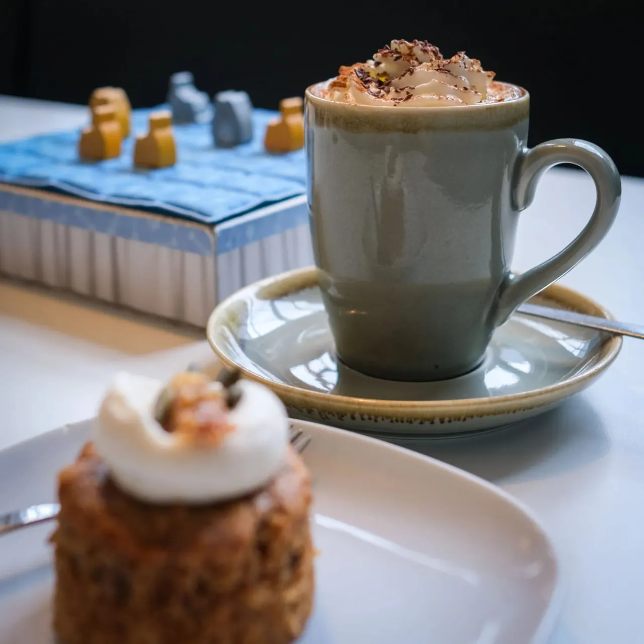 A mug of hot chocolate topped with whipped cream and chocolate shavings sits on a saucer, with pastries and cakes nearby on white plates.