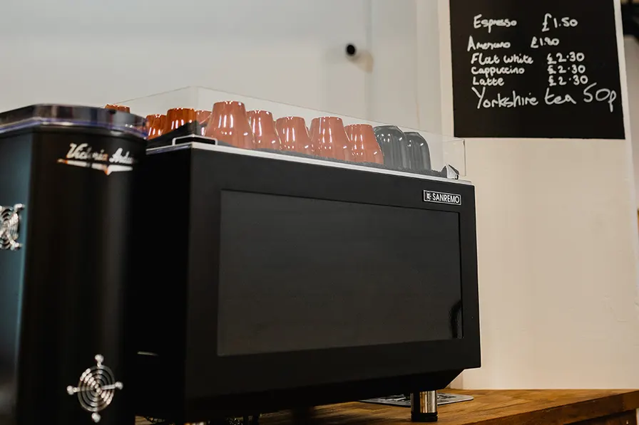 Espresso machine with cups on top and handwritten menu board displaying coffee prices above.