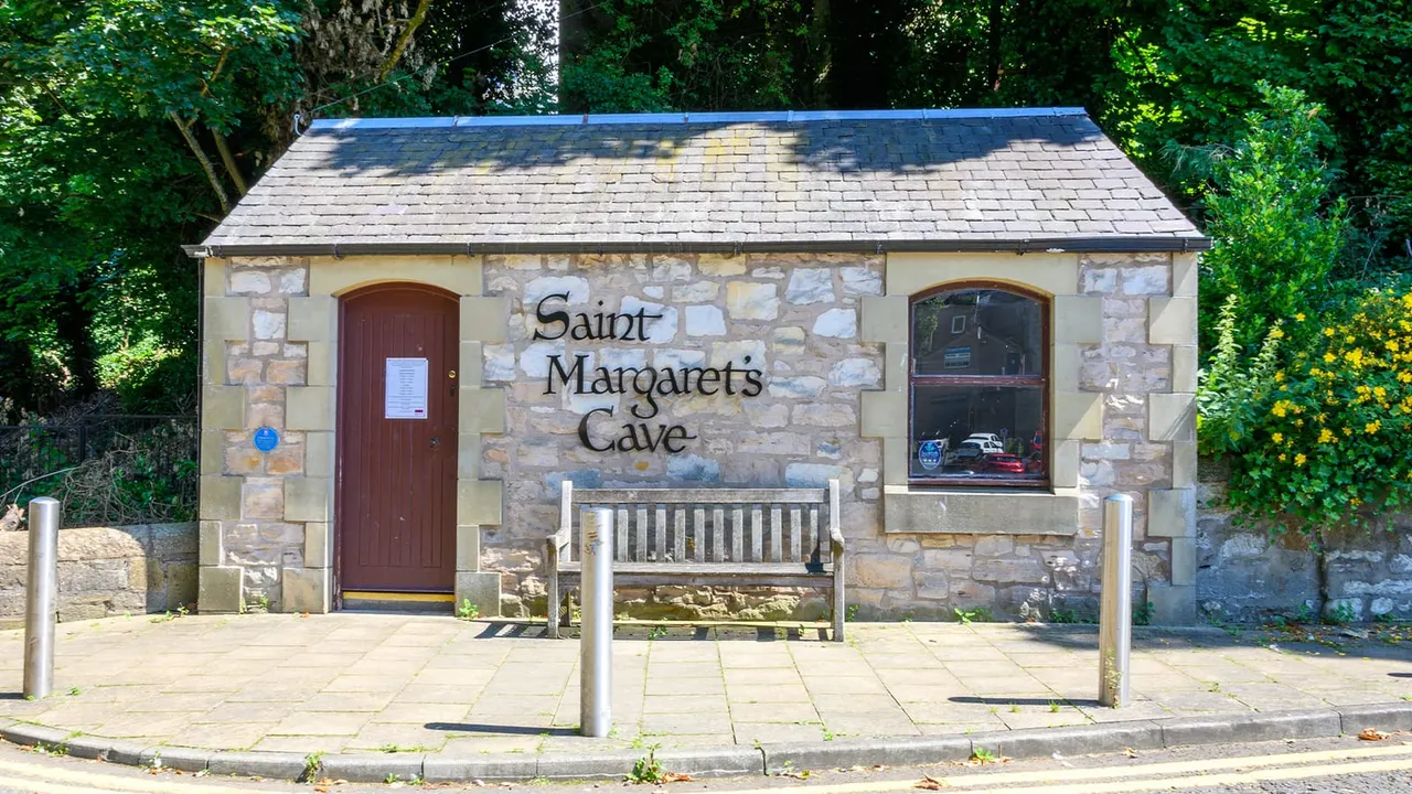 Stone building with red door and wooden bench, surrounded by greenery and bollards.