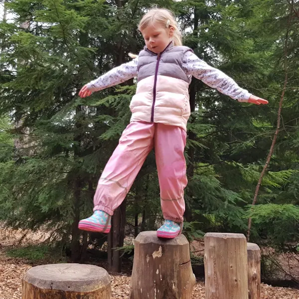 Young child balancing on wooden stumps with arms outstretched in a woodland adventure play area.