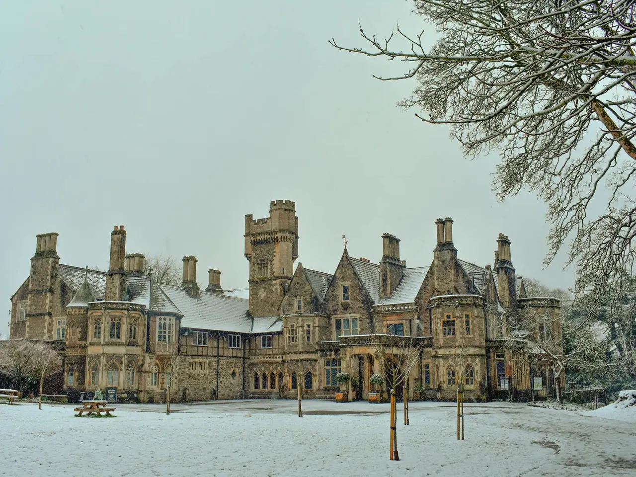 A grand Victorian stone mansion with multiple towers and chimneys stands in a snowy courtyard, framed by bare winter branches.