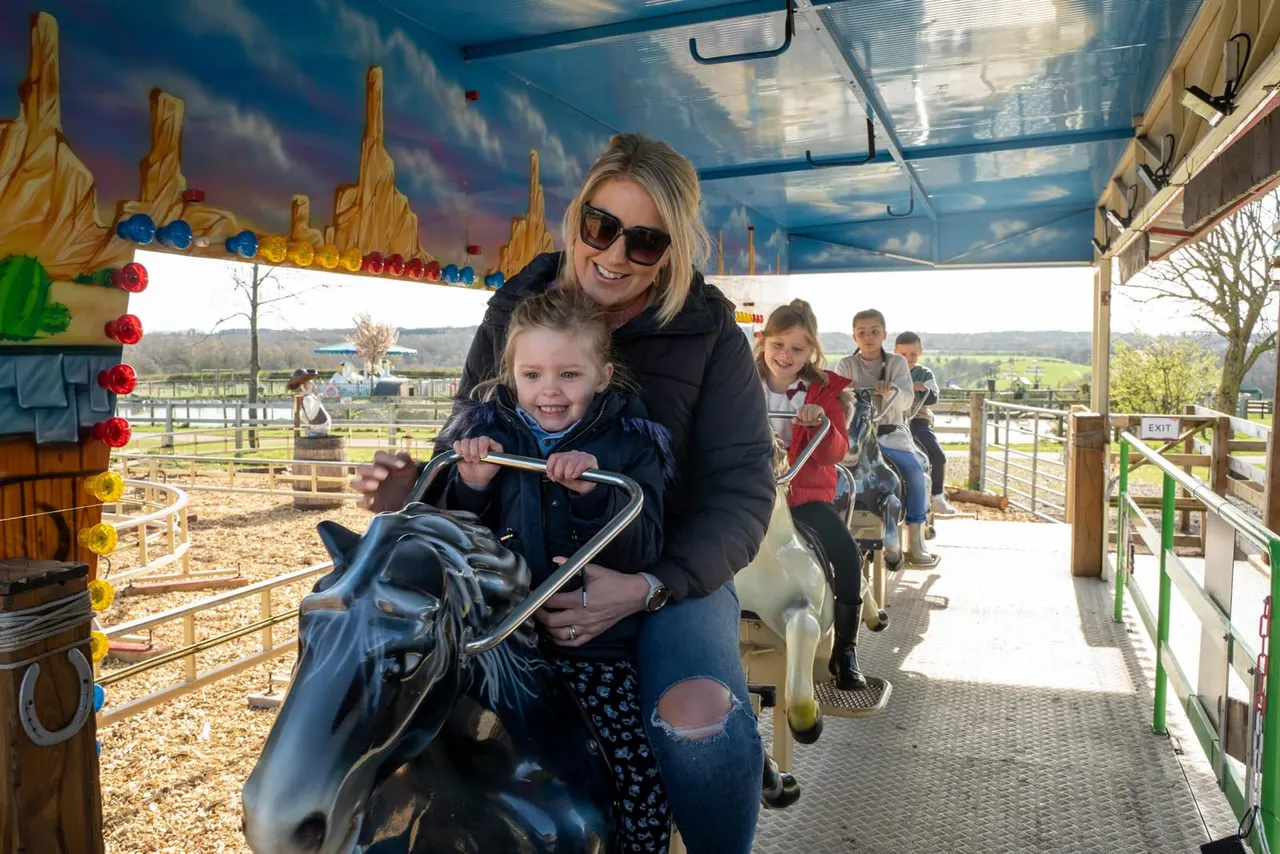Woman and young child on a themed ride vehicle with other children seated behind, passing decorated farm areas with countryside views.