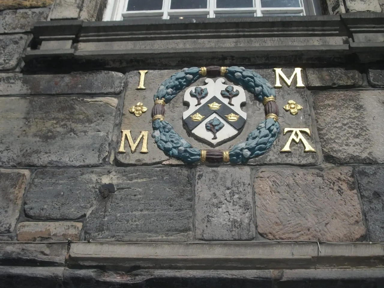Decorative stone plaque on historic building facade with heraldic coat of arms, gold lettering and blue wreath detail.