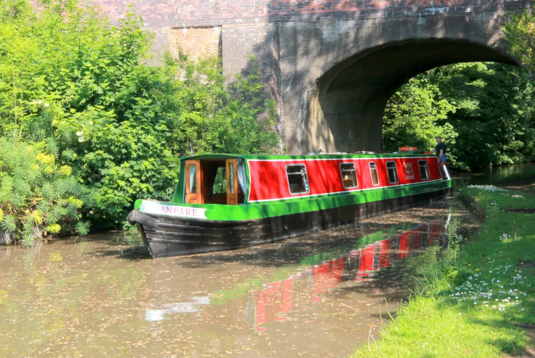 Red and green narrowboat moored on a canal beneath a stone arch bridge, surrounded by green vegetation.