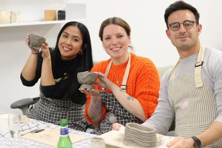 Three people wearing aprons hold up clay pieces they've made during a pottery workshop.